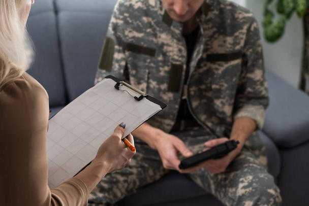 精神科医、ジェスチャー、兵士が心理学者、診療所のインテリアの前のソファに座っていると彼の考えを共有する感情的な軍人。PTSDと軍務. - 写真・画像