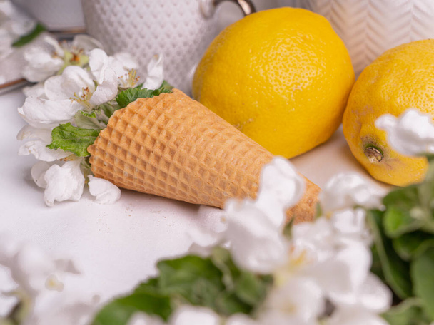 Set de té blanco con té en tazas con limones amarillos y una rama de manzana floreciente.AcercamientoHay conos de waffle en un platillo. - Foto, Imagen