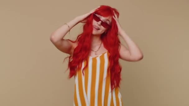 Funny joyful sincere redheadwoman in red sunglasses making playful silly facial expressions and grimacing, fooling around showing tongue. Young beautiful girl isolated alone on beige wall background - Footage, Video