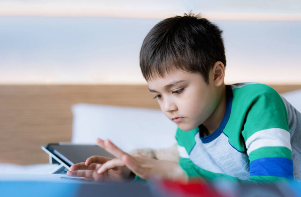 Cinematic portrait Kid playing games online on internet with friends in bed room, Young boy using tablet typing or chatting  at night, Childhood sitting on bed relaxing at home on weekend. - Photo, Image