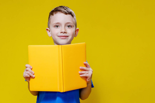 Little cheerful blond green-eyed boy 5-6 years old in a stylish blue T-shirt holding book and reading on yellow wall background, children's studio portrait. - Fotografie, Obrázek