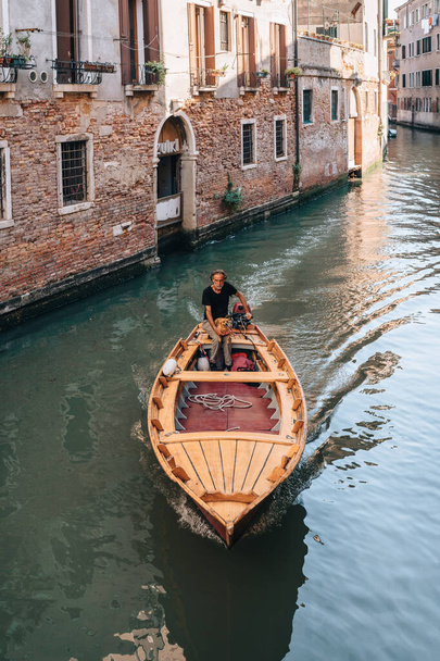 ヴェネツィア、イタリア- 2022年5月21日:犬がヴェネツィアの狭い運河で小さな木製のボートを運転している地元のイタリア人男性。ボートは市内の交通の主なモードです。. - 写真・画像