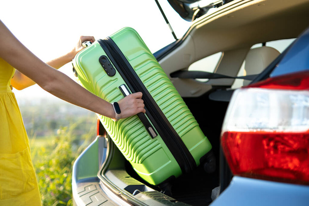 Close up of woman hands taking green suitcase from car trunk. Travel and vacations concept. - Фото, изображение
