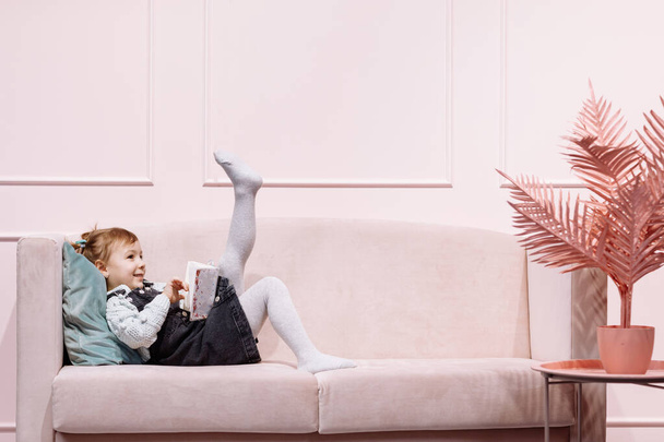 Little cute adorable girl is lying alone on pink sofa and reading book for childs in living room with decor - Foto, immagini