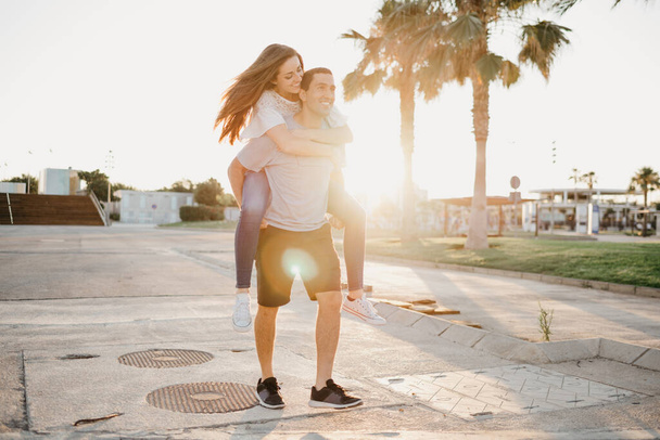 Een lachend brunette meisje rijdt 's avonds op haar Spaanse vriendje tussen palmen en zonnestralen in Spanje. Een paar toeristen op een date bij zonsondergang in Valencia. - Foto, afbeelding
