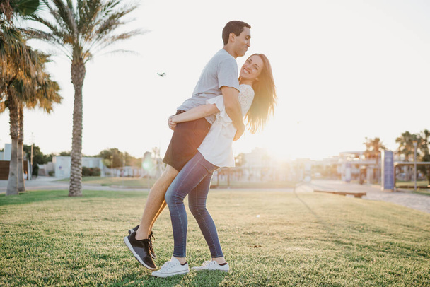 Une photo pleine longueur d'une fille brune heureuse qui soulève son petit ami hispanique entre les palmiers et la lumière du soleil en Espagne dans la soirée. Quelques touristes à un rendez-vous au coucher du soleil à Valence. - Photo, image