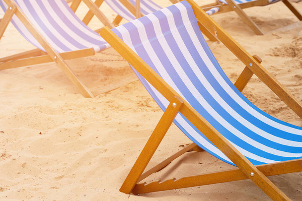 Сине-белые полосатые шезлонги, стоящие на песке на пляже, освещенном солнечными лучами - Фото, изображение