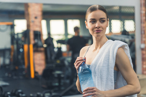 Μια γυμνασμένη γυναίκα με αθλητικά και μια πετσέτα στον ώμο της πίνει νερό στο γυμναστήριο. Υψηλής ποιότητας φωτογραφία - Φωτογραφία, εικόνα