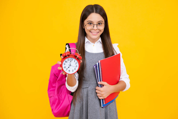 Шкільна дівчина-підліток перевіряє час. Школярка з будильником. Шкільний час. Щасливе обличчя, позитивні і усміхнені емоції школярки-підлітка ізольовані на жовтому
 - Фото, зображення