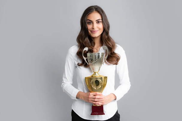Young happy woman holding a champion cup on gray background. Portrait of young woman with gold trophy cup. Successful employee holds winner cup. Award ceremony for winner. Best manager - Photo, Image