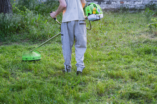 Der Kerl mäht, schneidet, schneidet Gras in einem zugewachsenen Garten, Gartenschneider oder Motorsense. Gartenarbeit, Gartenreinigung. Uns ist das egal... - Foto, Bild
