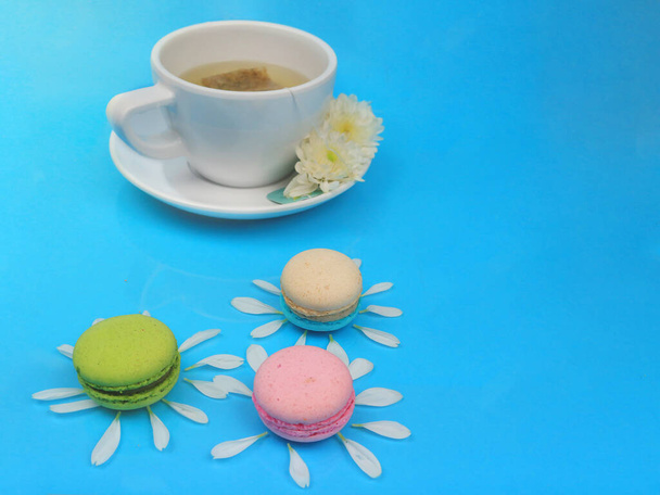 Schattige madeliefje macarons, kleurrijke Franse macarons met Chrysant bloemblaadje een kopje thee, heerlijk zoet dessert op blauwe achtergrond met copyspace, food achtergrond concept. - Foto, afbeelding