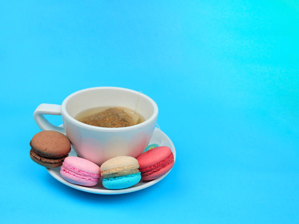 A cup of tea with cute colorful French macarons (macaroons) , delicious sweet dessert on blue background, lovely food background concept. - Foto, afbeelding