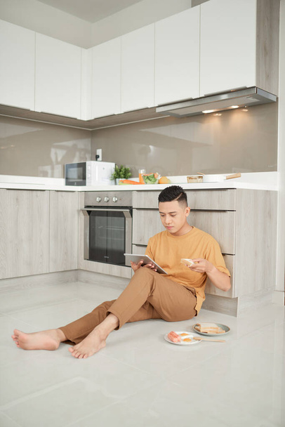 デジタルタブレットを使用してハンサムな若い男とキッチンフロアに座って朝食を持っています。 - 写真・画像