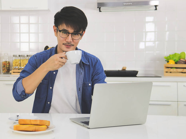 Retrato de un joven asiático guapo con anteojos sentados en la mesa de la cocina con computadora portátil y pan tostado en un plato, sonriendo sosteniendo una taza de café, mirando a la cámara - Foto, imagen