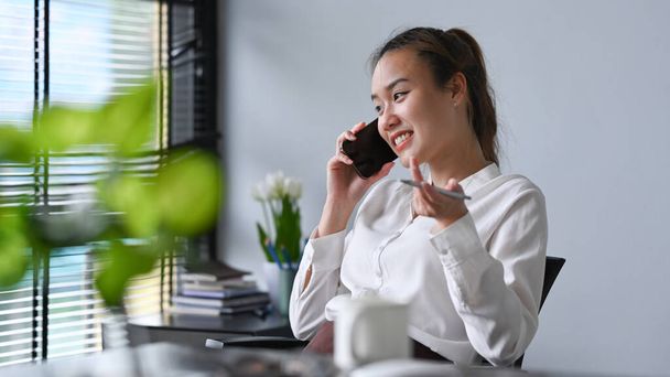 Επαγγελματική νεαρή επιχειρηματίας που έχει κινητό τηλέφωνο συνομιλία, ενώ κάθεται στο σύγχρονο χώρο εργασίας. - Φωτογραφία, εικόνα