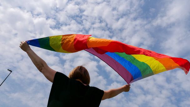 LMBTQIA hónap büszkeség, szivárvány béke zászló ellen kék ég felhők egy napsütéses napon. Tartsd a nőt a kezedben és ünnepeld a biszexualitás napját vagy a nemzeti kijövetel napját. - Fotó, kép