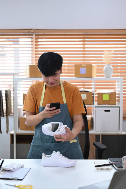 Młody człowiek pracujący w biurze domowym i robiący zdjęcia do butów ze smartfonem na pocztę do sprzedaży online w Internecie. - Zdjęcie, obraz