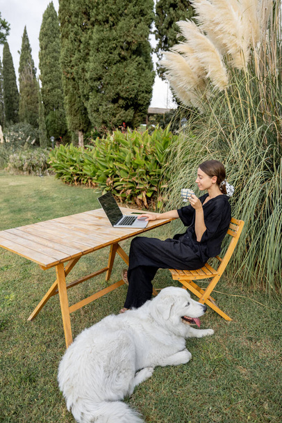 Meisje strelen Maremmano-Abruzzese Sheepdog en met behulp van laptop computer aan houten tafel op groene mediterrane gazon. Begrip weekend, rust en vakantie. Idee van vriendschap van mens en huisdier - Foto, afbeelding
