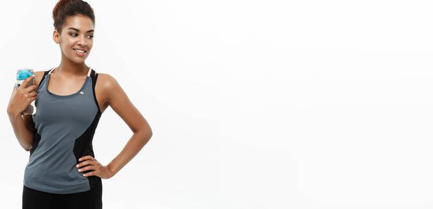 Υγιείς και Fitness έννοια - όμορφη αφρικανική αμερικανική κορίτσι στον αθλητισμό ρούχα μπουκάλι νερό εκμετάλλευση μετά την προπόνηση. Απομονωμένα σε φόντο λευκό στούντιο. - Φωτογραφία, εικόνα