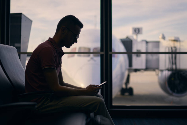 Silhouette of young man waiting while waiting for flight. Traveler using phone in airport terminal. - 写真・画像