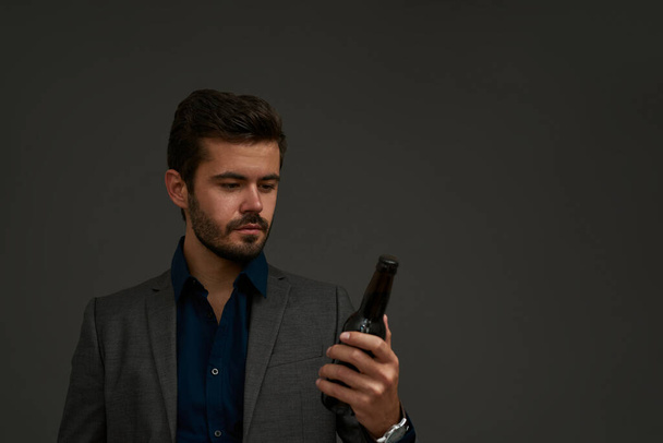 Concentrated caucasian brewer entrepreneur looking at beer bottle. Bearded man wearing formal wear. Concept of brewing business. Isolated on grey background. Studio shoot. Copy space - Photo, Image