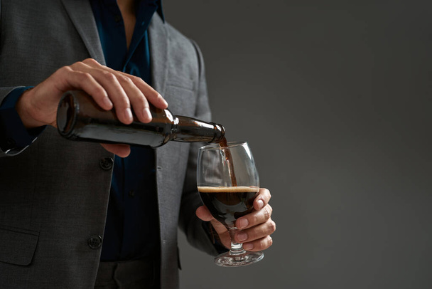Cropped of brewer entrepreneur pouring beer in glass from bottle. Man wearing suit. Concept of brewing business. Modern successful male lifestyle. Isolated on grey background. Studio shoot. Copy space - Foto, Bild