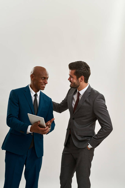 Black businessman show somes on digital tablet to caucasian colleague. Smiling multiracial men look at each other. Modern successful male lifestyle. Teamwork. White background in studio. Copy space - Photo, image