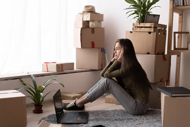 Atrakcyjna młoda kobieta porusza się, siedząc wśród kartonów, korzystając z laptopa i uśmiechniętego relaksującego, poruszającego się konceptu dnia - Zdjęcie, obraz