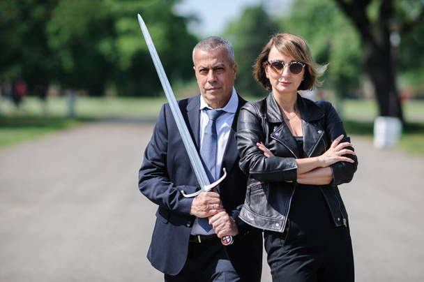 Portrait of handsome businessman model man in a modern casual suit protect woman love with a large medieval sword. Modern fashion street style concept. Elegant urban warrior couple - 写真・画像