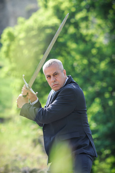 Portrait of handsome businessman model man in a modern casual suit with a large medieval sword. Modern fashion street style concept. Elegant urban warrior. Corporate and money security - Foto, afbeelding