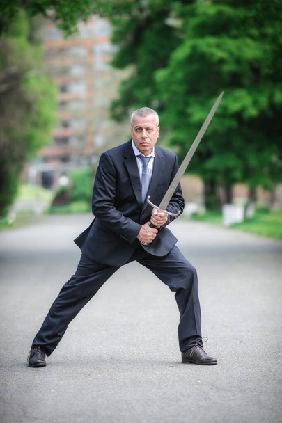 Portrait of handsome businessman model man in a modern casual suit with a large medieval sword. Modern fashion street style concept. Elegant urban warrior. Corporate and money security - Фото, зображення