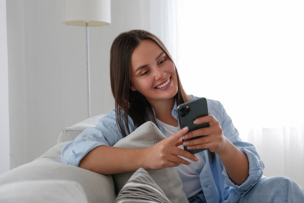 Portrait of young brunette woman wearing mom jeans sitting on the couch texting and smiling. Joyful female having fun video phone call at home. Background, copy space, close up. - Photo, image