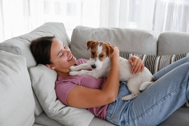 Portrait of young beautiful woman lying on the couch with her adorable wire haired Jack Russel terrier puppy. Loving girl with rough coated pup having fun. Background, close up, copy space. - 写真・画像