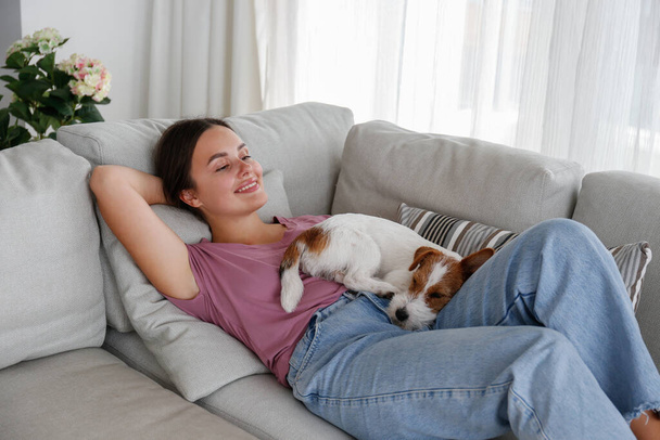 Portrait of young beautiful woman lying on the couch with her adorable wire haired Jack Russel terrier puppy. Loving girl with rough coated pup having fun. Background, close up, copy space. - Fotó, kép