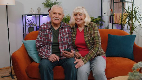 Mayores ancianos familia pareja abuelos hombre mujer en casa sala de estar y sentado en el sofá. Abuela madura abuelo trabaja en smartphone, envía mensajes, hace compras en línea - Foto, imagen
