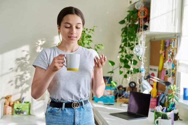 Teenage girl 14, 15 years old holding a mug, standing at home near sunny window, table. Beautiful attractive female in home interior resting smiling. Adolescence, lifestyle, young people concept - Photo, Image