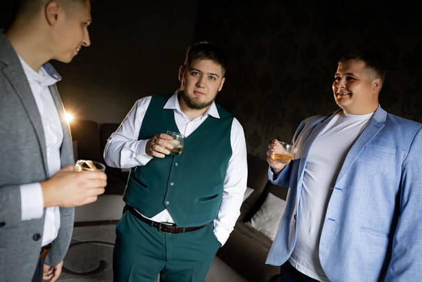 Three men with glasses of whiskey stand near the window in the apartment. Natural daylight. Groom's Gathering - Foto, Bild