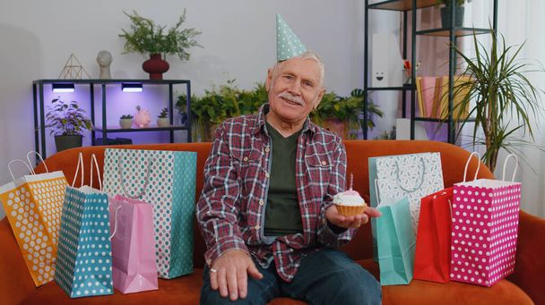 Glücklicher älterer Herr mit festlichem Geburtstagshut hält Cupcake in der Hand und gratuliert mit brennender Kerze auf Kuchen. Reifer Großvater feiert Party auf Couch mit Geschenken zu Hause - Foto, Bild