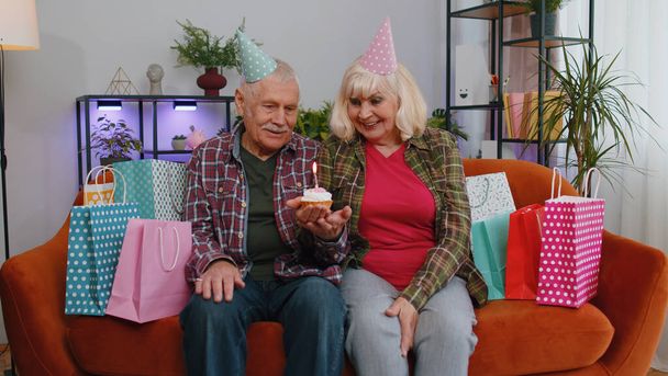 Χαρούμενη ηλικιωμένη οικογένεια ζευγάρι παππούδες άντρας γυναίκα φοράει εορταστικό καπέλο καπέλο κρατήσει cupcake κάνει ευχή γιορτάζει επέτειο φυσώντας κερί καύση στην τούρτα. Γιαγιά παππούς με δώρα στο σπίτι - Φωτογραφία, εικόνα