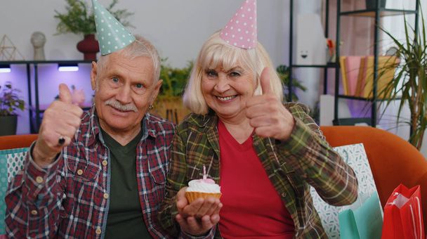 Happy senior family couple grandparents man woman wears festive birthday hat hold cupcake makes wish celebrating anniversary blowing burning candle on cake. Grandmother grandfather with gifts at home - Photo, Image