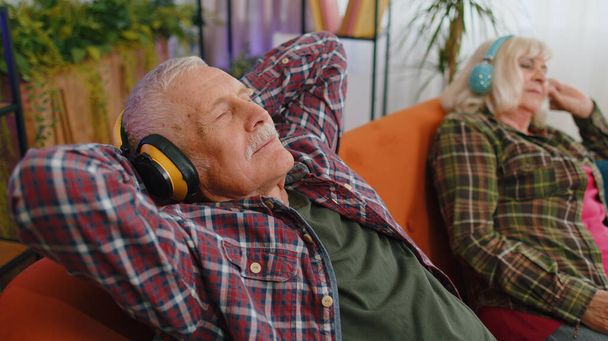Iloinen vanhempi vanhempi perhe pari isovanhemmat mies nainen käyttää kuulokkeet kuunnella musiikkia rentouttavaa istuu sohvalla kotona viikonloppuna. Kypsä isoäiti isoisä lepää yhdessä sohvalla. - Valokuva, kuva