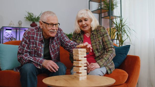 Stara starsza starsza rodzina para dziadków mężczyzna kobieta gra w bloki budować grę. Dojrzałe babcia dziadek spędzać czas razem grać w gry planszowe hobby z drewnianą wieżą na kanapie w domu - Zdjęcie, obraz