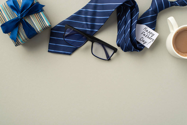 Vatertagskonzept. Foto von oben von Geschenkbox mit Schleife blaue Krawatte Gläser Postkarte und Tasse Kaffee auf isolierten pastellgrauen Hintergrund mit Copyspace - Foto, Bild