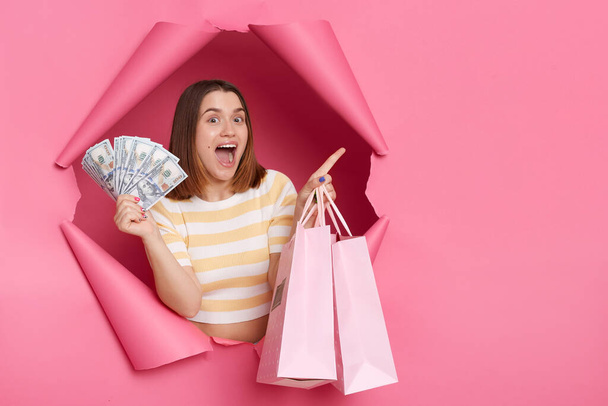 Portrait of excited surprised attractive woman wearing striped shirt looking through breakthrough of pink background, pointing finger aside, holding fan of dollars and shopping bags. - Фото, зображення