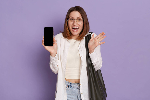 Excited optimistic woman with black bag wearing white shirt and jeans standing with cell phone, showing empty display with space for advertisement, posing isolated over purple background. - Φωτογραφία, εικόνα