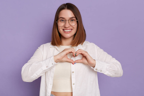 Portrait of smiling lovely woman wearing white shirt posing isolated over purple background, showing heart shape with hands, expressing romantic feelings. - Fotó, kép