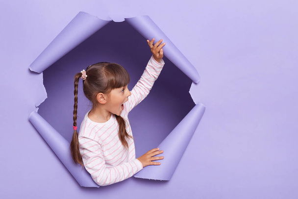 Horizontal shot of cute charming little girl with braids wearing striped shirt posing in torn paper wall, looking away with amazed expression, copy space for advertisement or promotion. - Photo, Image