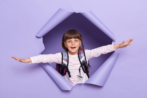 Portrait of happy cute little girl with pigtails wearing striped shirt with backpack raised arms pretending she is flying, raised arms and looking at camera. - Photo, Image