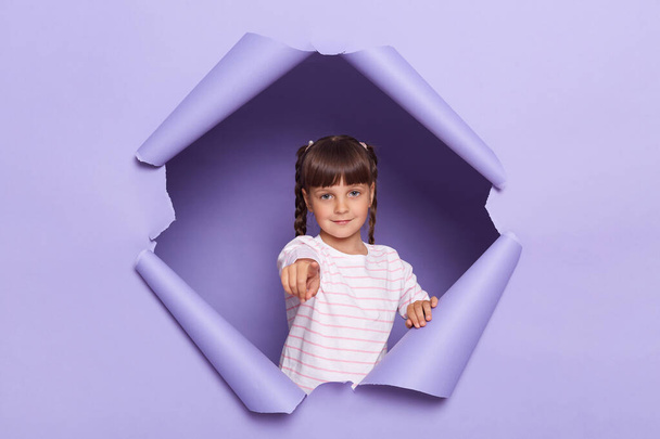 Portrait of serious little girl with pigtails wearing striped shirt breaks through purple paper background, female kid pointing finger and camera, choosing you, showing her choice. - Foto, afbeelding